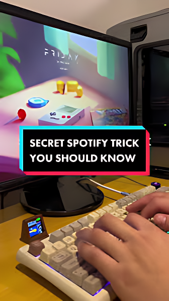 a close-up photograph showing someone's hands typing on a mechanical keyboard in front of a computer monitor. The monitor displays a colorful, cartoon-style illustration with playful objects on a table. In the center of the image, bold white text on a highlighted background reads: "SECRET SPOTIFY TRICK YOU SHOULD KNOW," suggesting the content relates to tips or hidden features of Spotify. The setting appears cozy and tech-oriented.